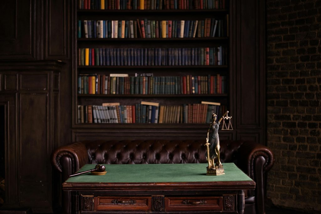 A classic study room with shelves of books, a gavel, and Lady Justice figurine on a green table.